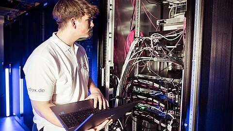Ein Techniker im weißen Poloshirt arbeitet in einem Rechenzentrum an einem Laptop vor einem Server-Rack voller Kabel und Netzwerkgeräte.