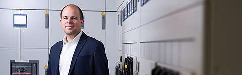 Ein Mann in einem dunklen Anzug und weißem Hemd steht in einem modernen Kontrollraum mit Schalttafeln und Monitoren im Hintergrund und blickt selbstbewusst in die Kamera.