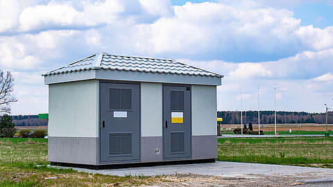 Ein kleines grau-weißes Versorgungsgebäude mit einem Metalldach steht auf einer Betonplatte in einem grasbewachsenen Gebiet mit Feldern und Bäumen im Hintergrund unter einem teilweise bewölkten Himmel.
