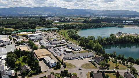 Luftaufnahme eines Industriegebiets mit Lagerhallen und Parkplätzen, umgeben von Grünflächen, zwei großen blauen Seen und entfernten Bergen unter einem teilweise bewölkten Himmel.