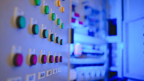 Nahaufnahme eines Bedienfelds mit Reihen bunter Knöpfe und Schalter, jeder beschriftet, vor einem unscharfen, blau beleuchteten Hintergrund elektrischer oder industrieller Geräte.