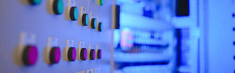 Nahaufnahme eines Bedienfelds mit runden Tasten in verschiedenen Farben vor einem unscharfen Hintergrund aus elektrischen oder industriellen Geräten, beleuchtet von blauem Licht.