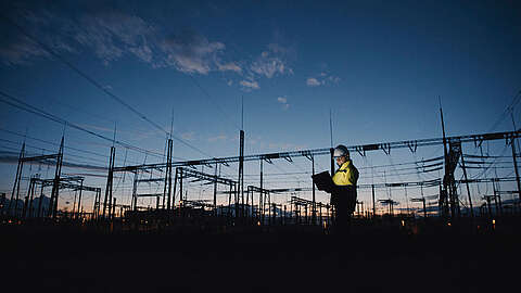 Ein Arbeiter, der einen Schutzhelm und eine Warnweste trägt, arbeitet in der Abenddämmerung an einem Laptop in einem Umspannwerk, wobei sich die Stromleitungen gegen den Abendhimmel abzeichnen.