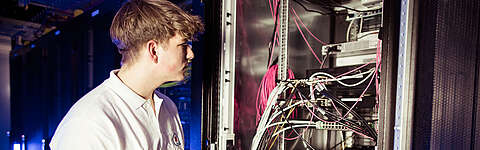 Eine Person in einem weißen Polohemd begutachtet einen Serverschrank mit Netzwerkkabeln und elektronischen Geräten in einem Rechenzentrum.