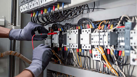 Eine Person mit grauen Handschuhen prüft mit einem Multimeter die elektrischen Verbindungen in einem Schaltschrank mit Drähten, Schaltern und Leistungsschaltern.