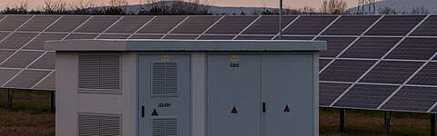Ein weißer Stromversorgungskasten steht vor Reihen von Solarmodulen auf einem offenen Feld, mit fernen Hügeln und einem Sonnenuntergangshimmel im Hintergrund.